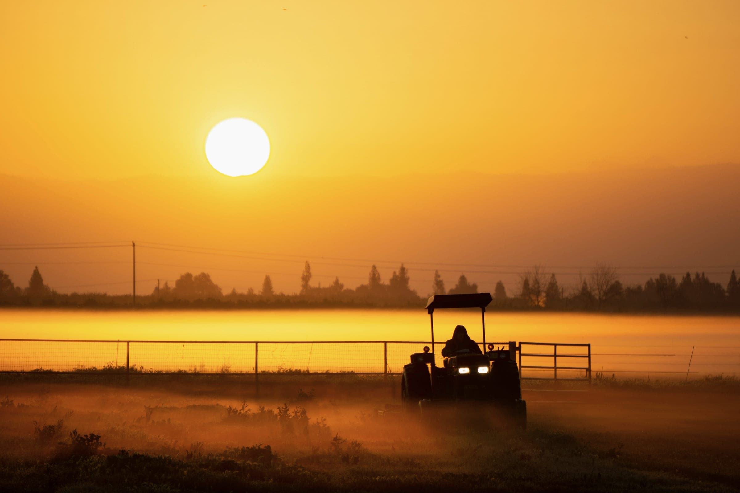 A tractor silhouetted against a low orange sun and rising mist in a farm field at dawn.