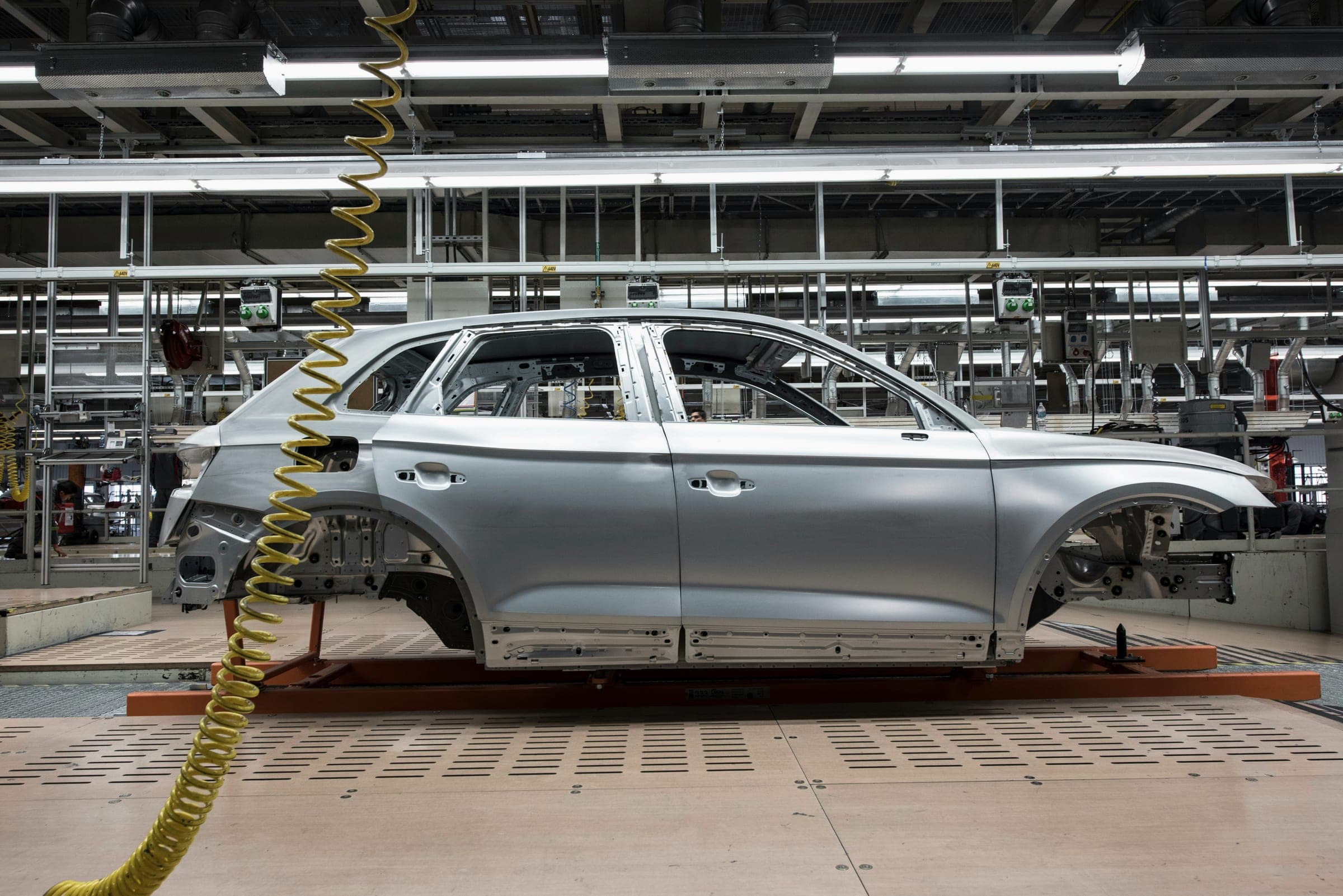 A bare silver car body shell on an assembly line carrier under rows of factory lights.