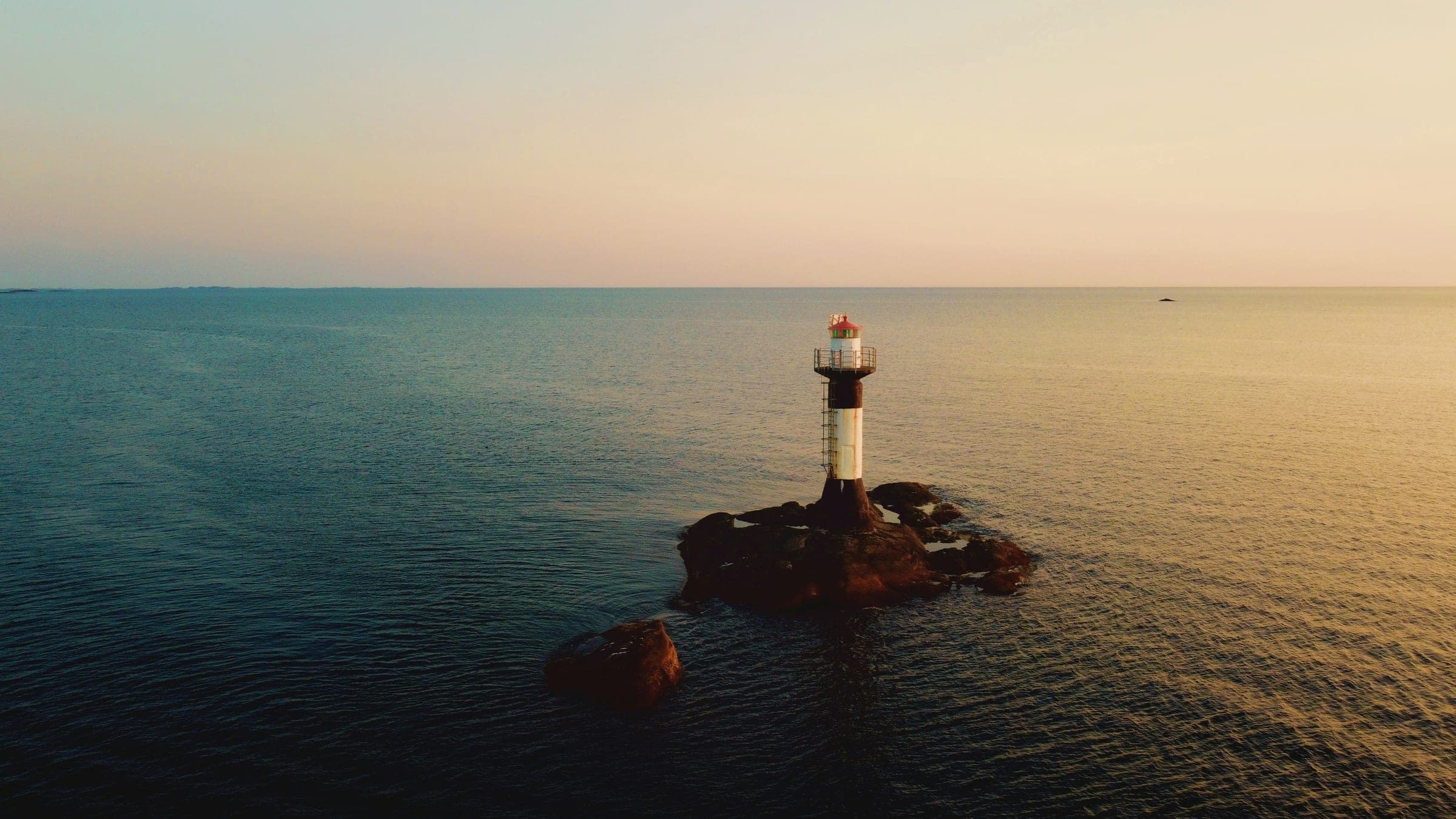 A small lighthouse on a rock in a calm sea under a soft peach and gold dusk sky.