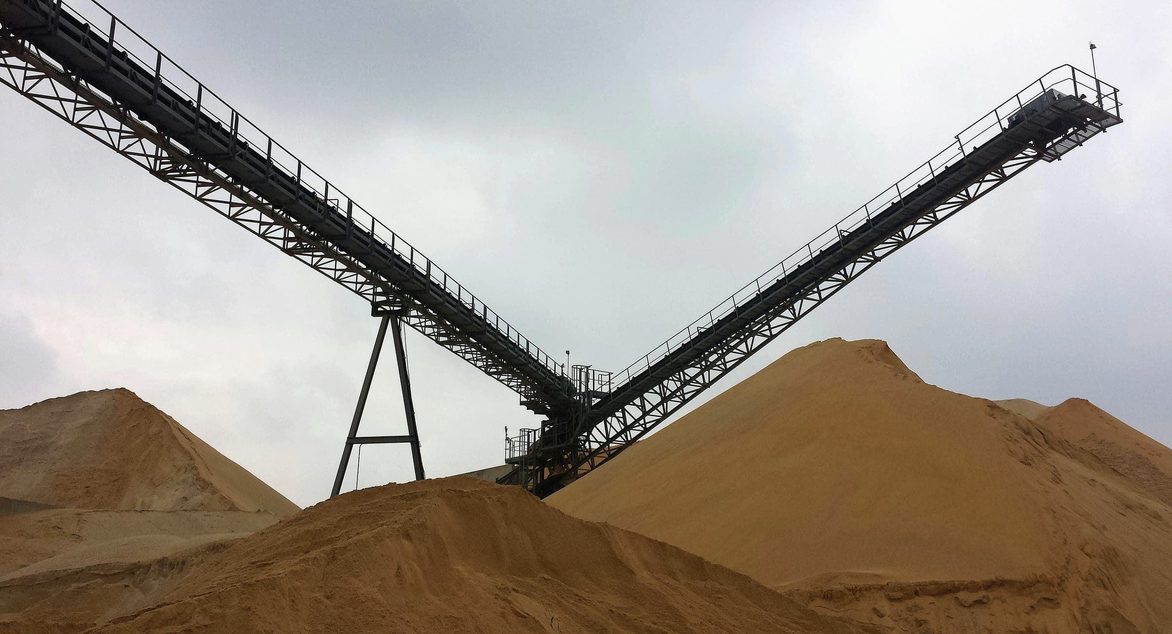 Two conveyor gantries crossing above large piles of sand under an overcast sky.