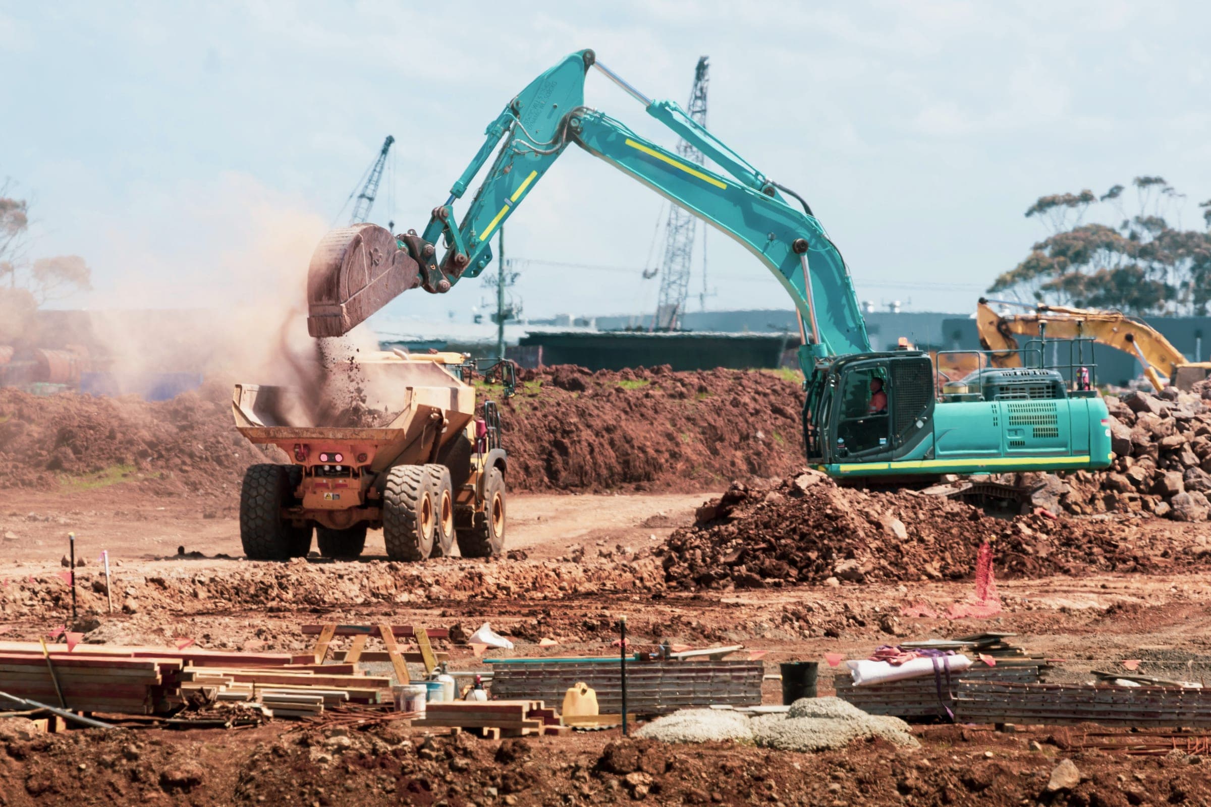A teal excavator loading soil into a dump truck at an earthworks site, with cranes in the distance.