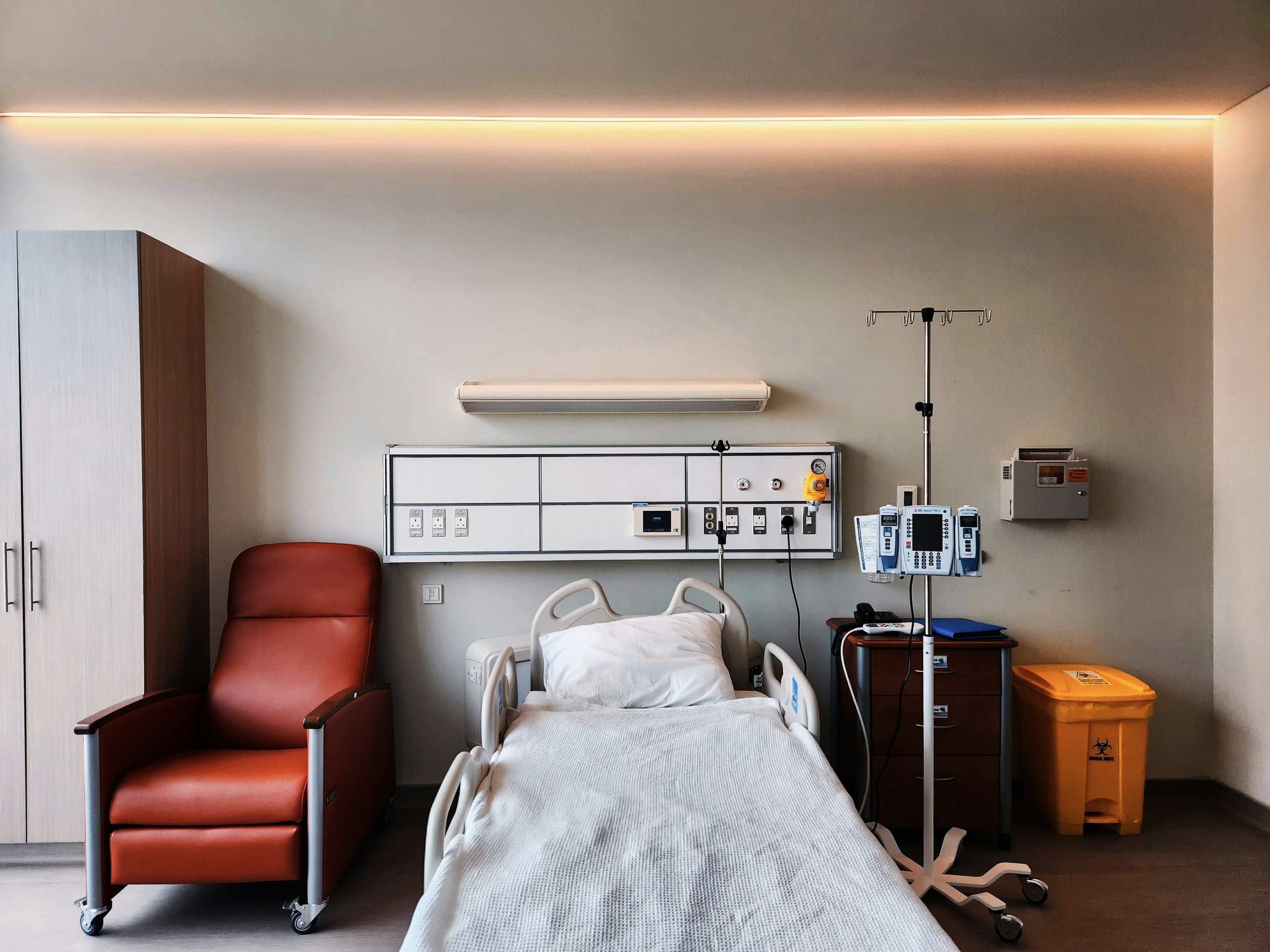 An empty modern hospital room with a made bed, an orange recliner, an IV stand and a warm strip of ceiling light.