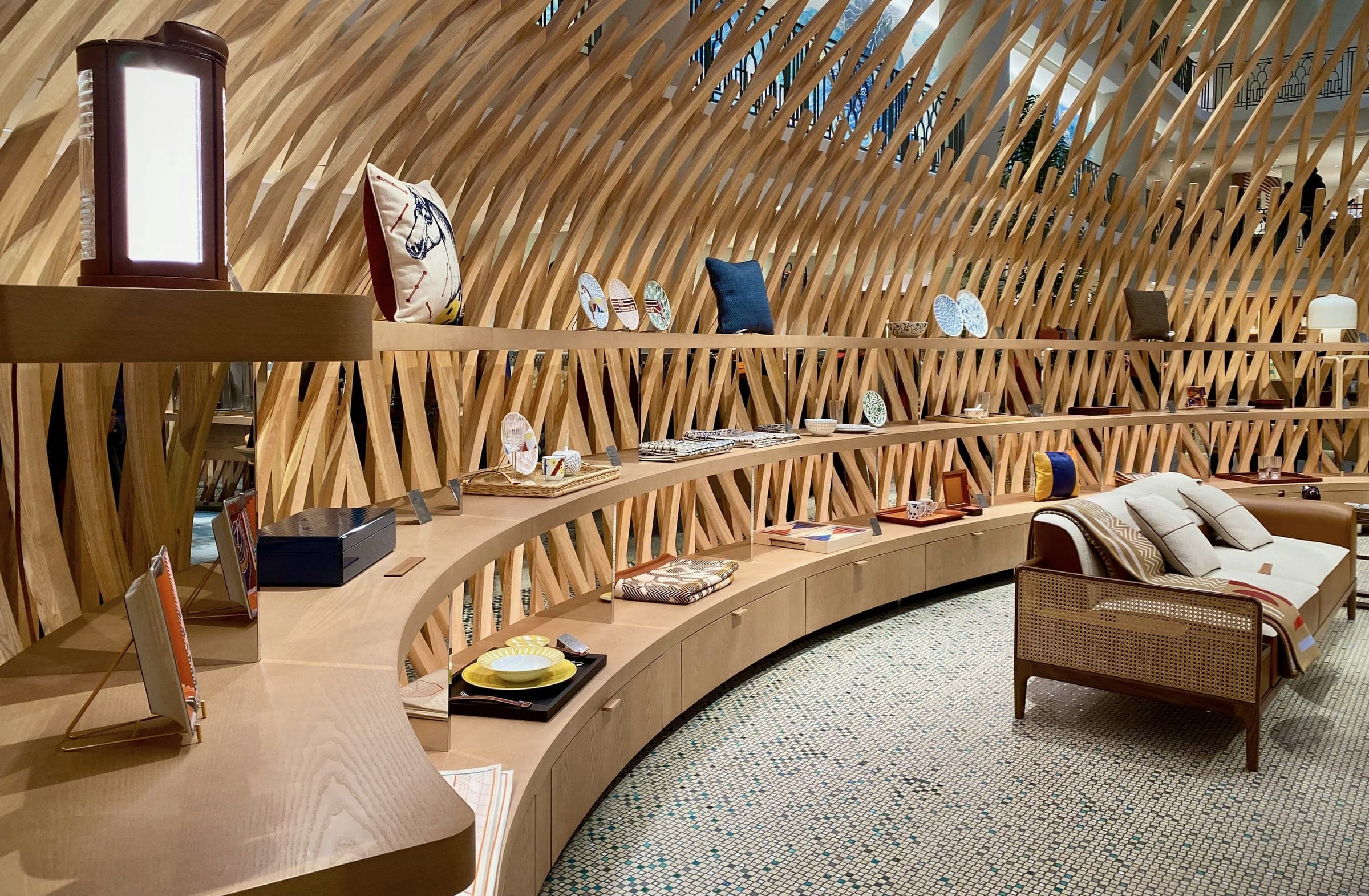A curved wooden showroom display with cushions, plates and trays beside a cane-backed sofa under a lattice of timber slats.