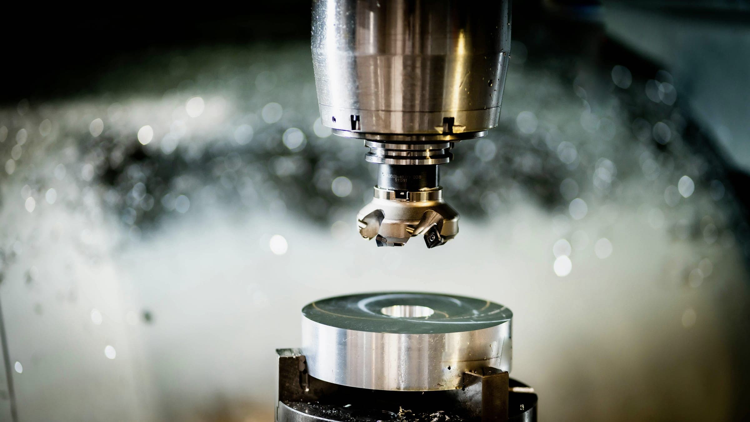 A milling cutter above a polished metal disc on a CNC machine, with coolant spray blurred behind it.