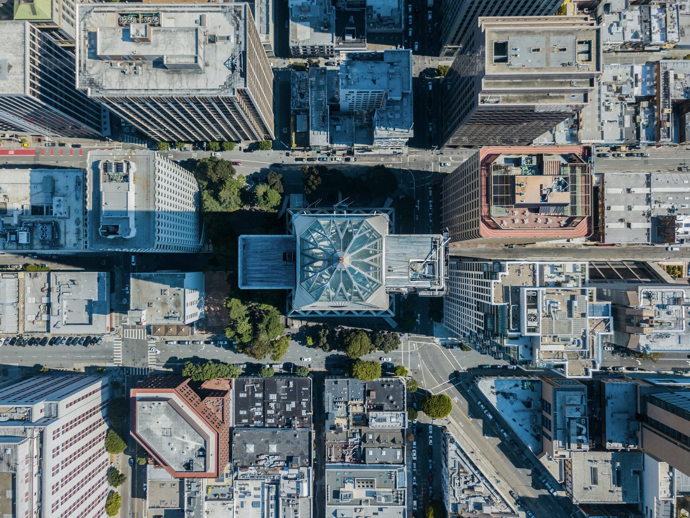 Top-down aerial view of a dense city grid of commercial buildings, rooftops and tree-lined streets in cool blue light.