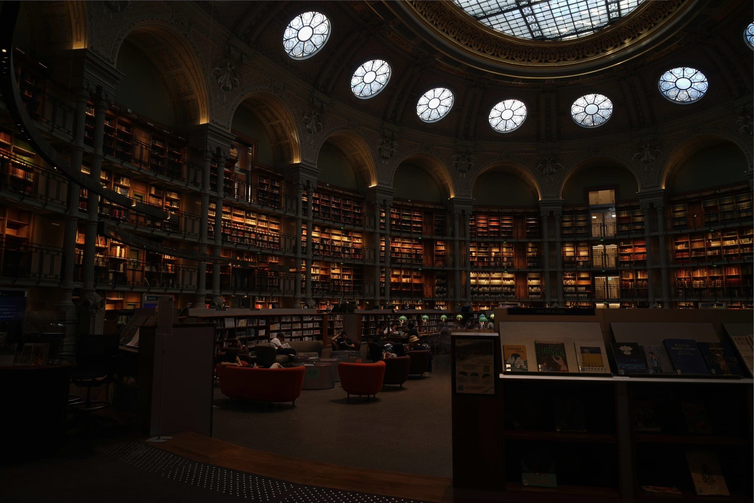 A dimly lit domed library reading room with round skylights, tiers of bookshelves and a few red armchairs.