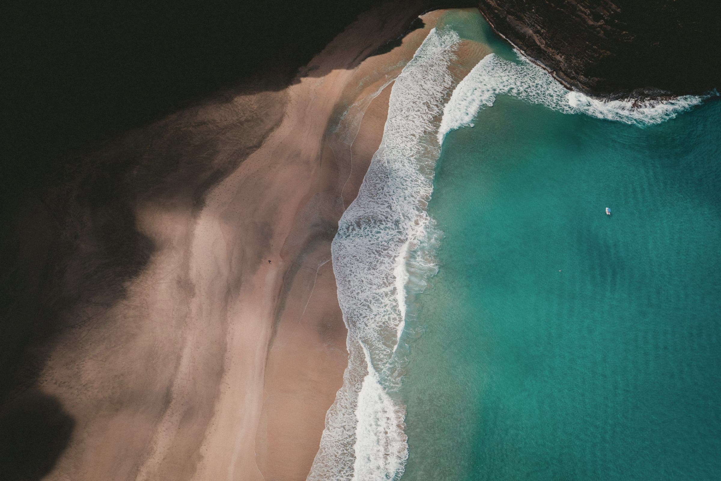 Aerial view of a calm turquoise sea washing onto a pale sand beach beside a dark rocky headland.