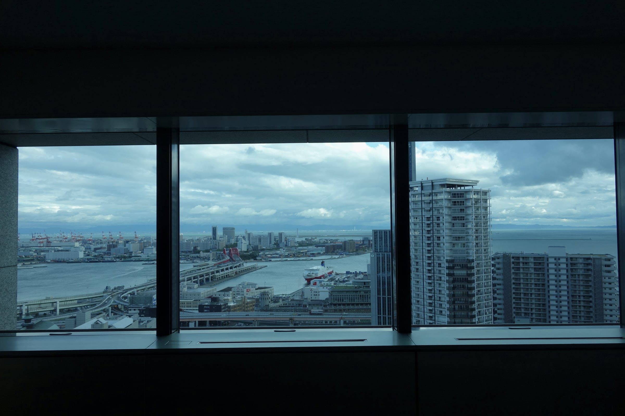 A dark office window framing a high-floor view of a harbor, port cranes and apartment towers under gray clouds.