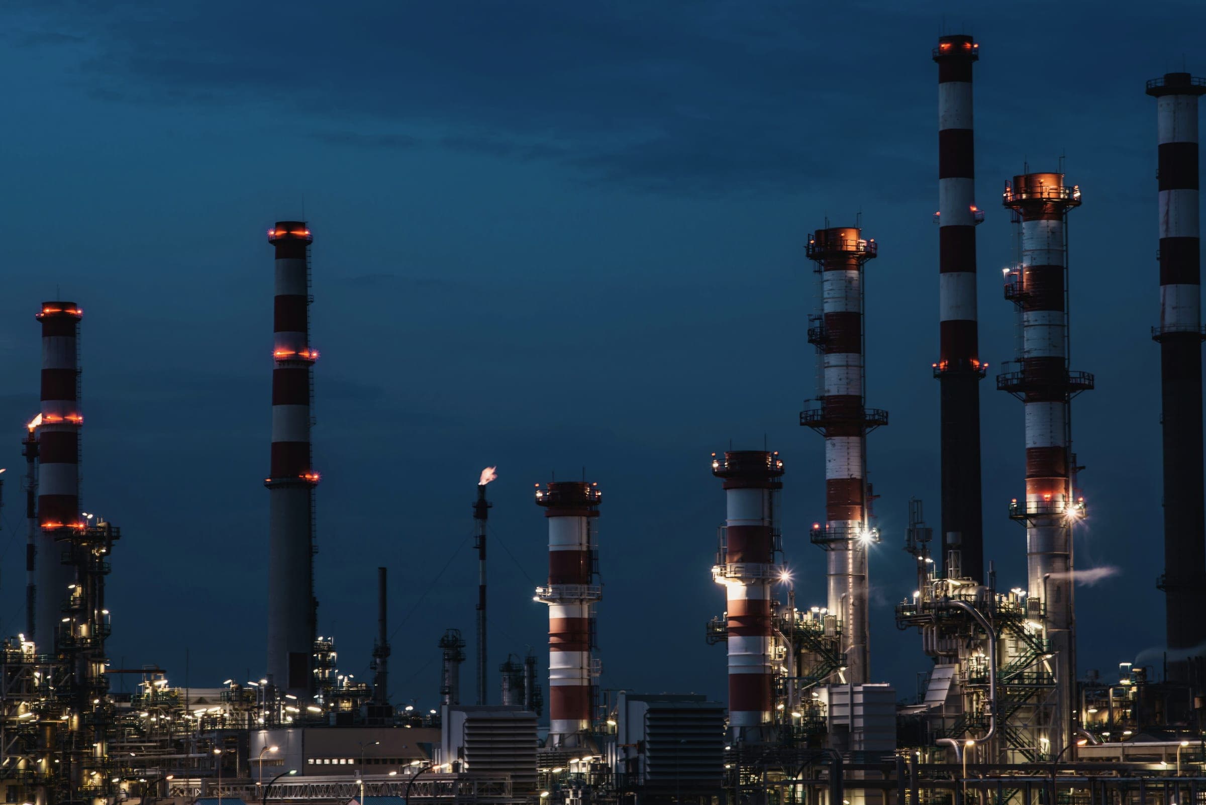 Striped refinery stacks and lit process towers against a deep blue evening sky.
