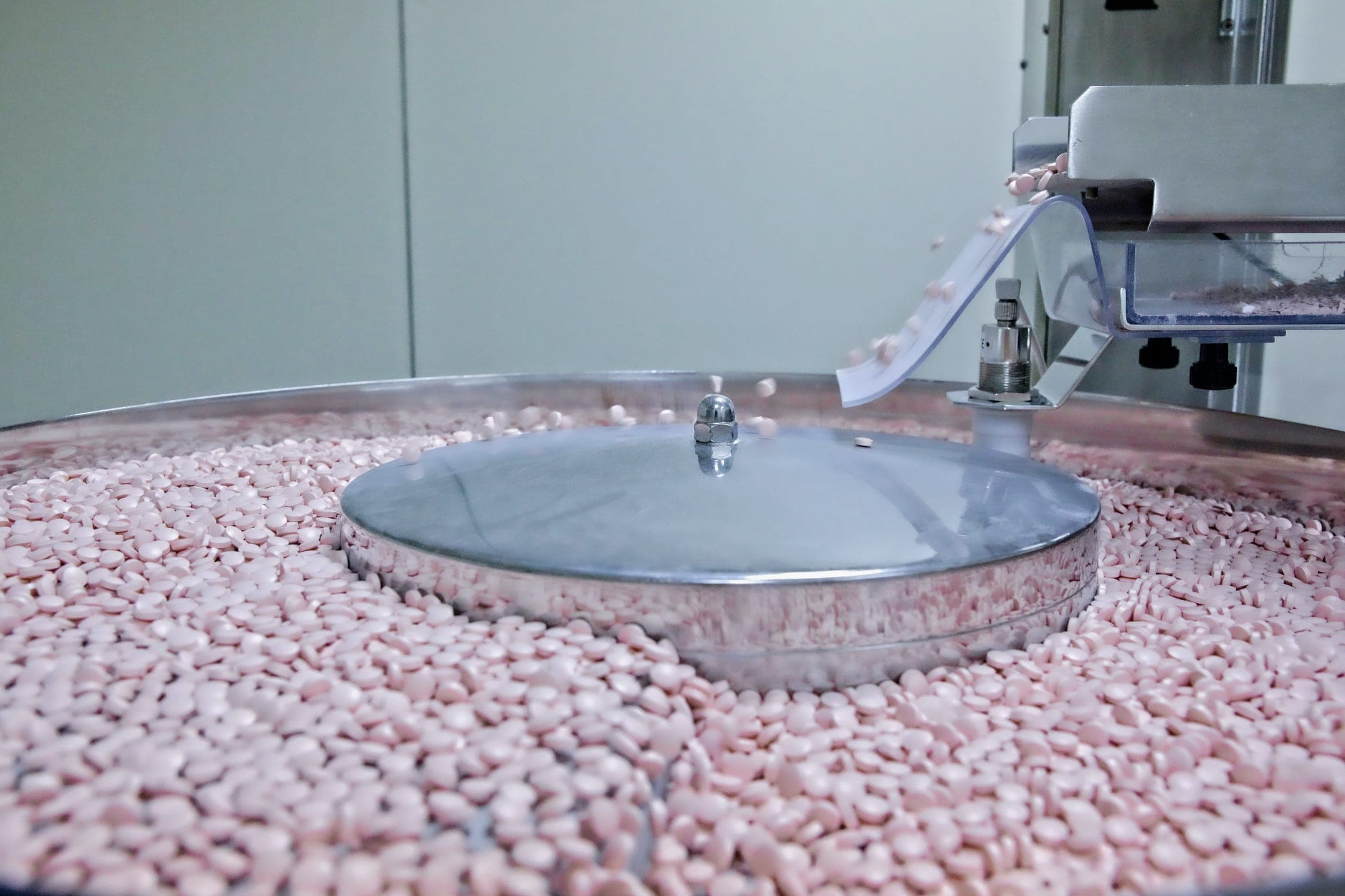 Pink tablets tumbling in a stainless steel coating drum at a pharmaceutical production machine.