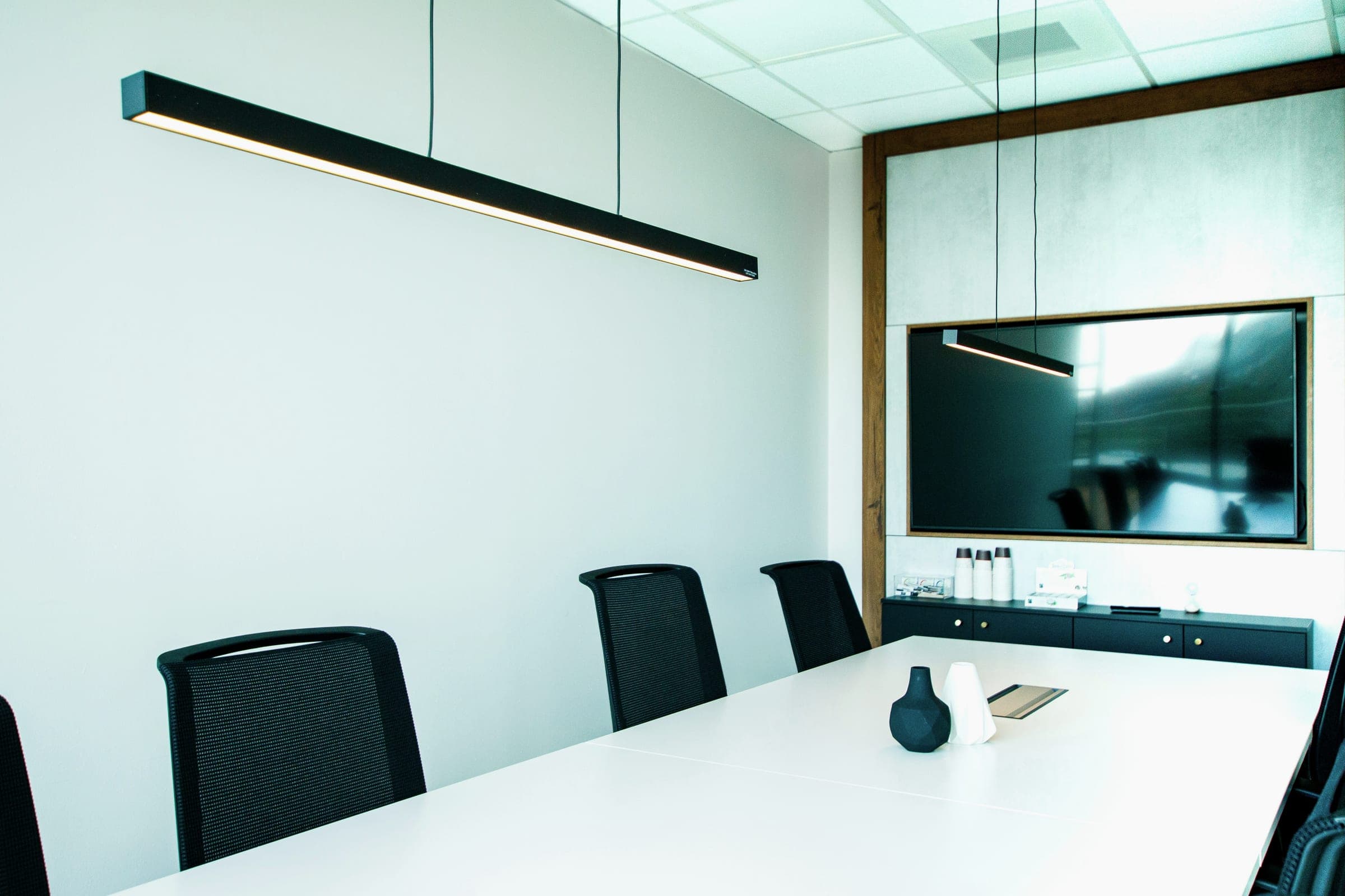 An empty modern meeting room with a white table, black mesh chairs, a wall display and linear pendant lights.