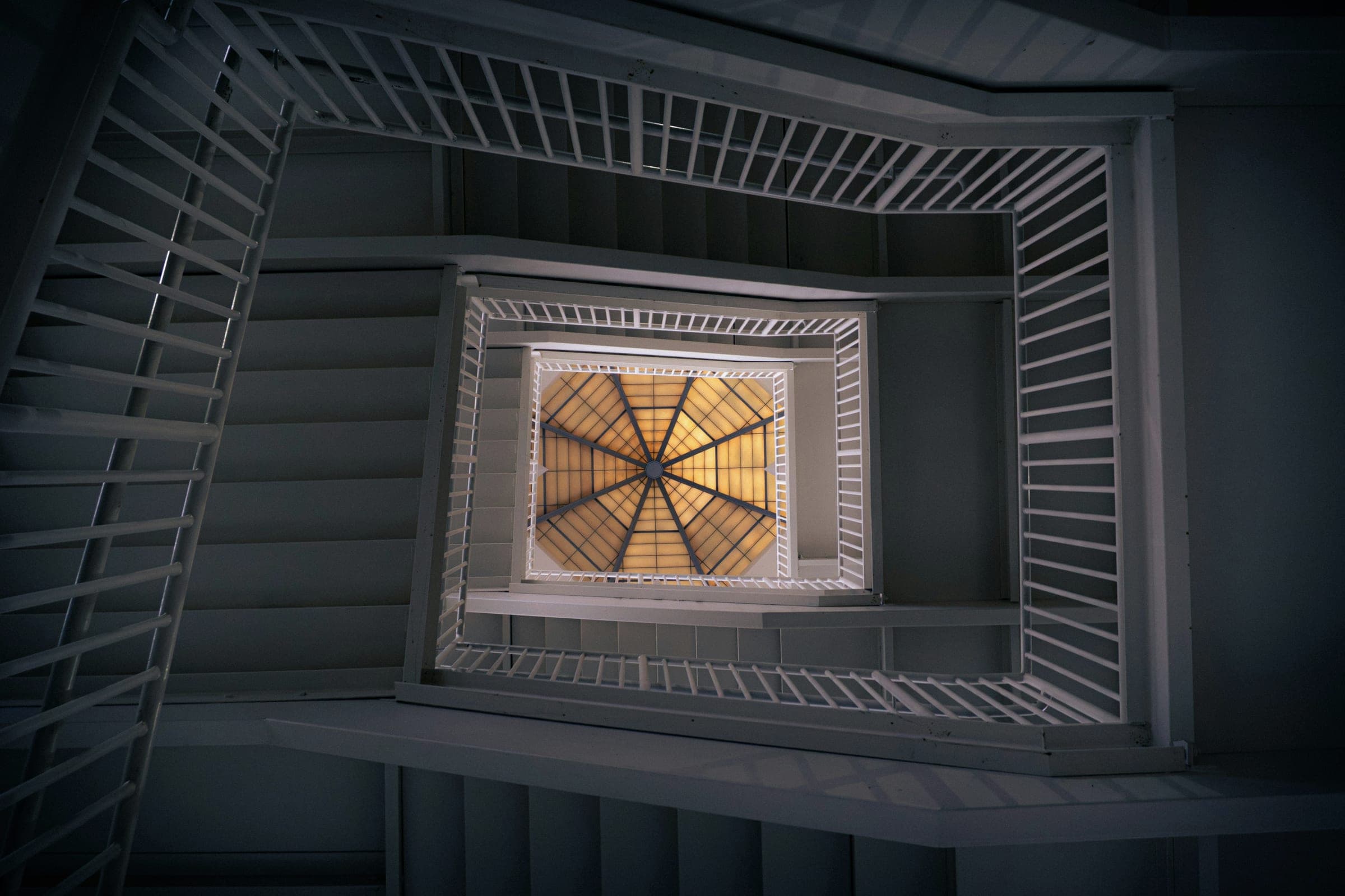 Looking straight up a square stairwell with white balustrades towards an octagonal, amber-lit skylight.