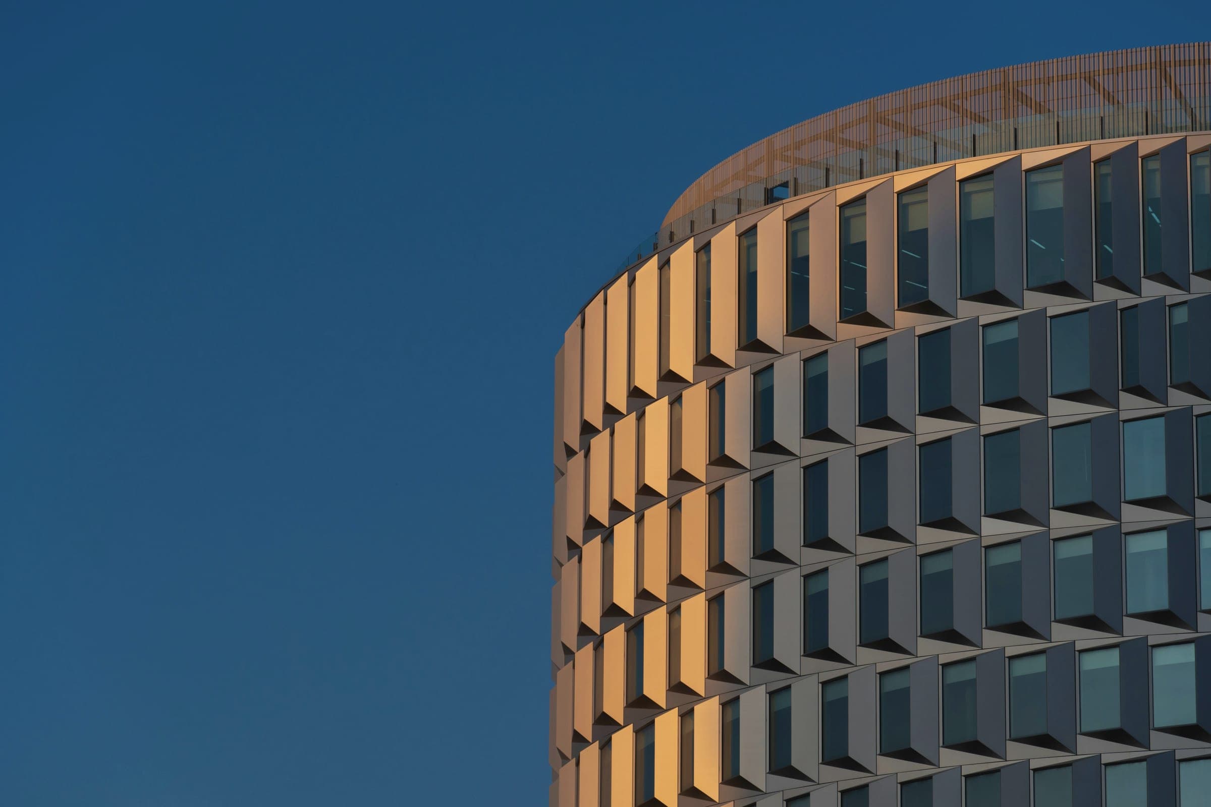 Curved office tower with warm, sunlit angled facade panels rising against a deep blue dusk sky.