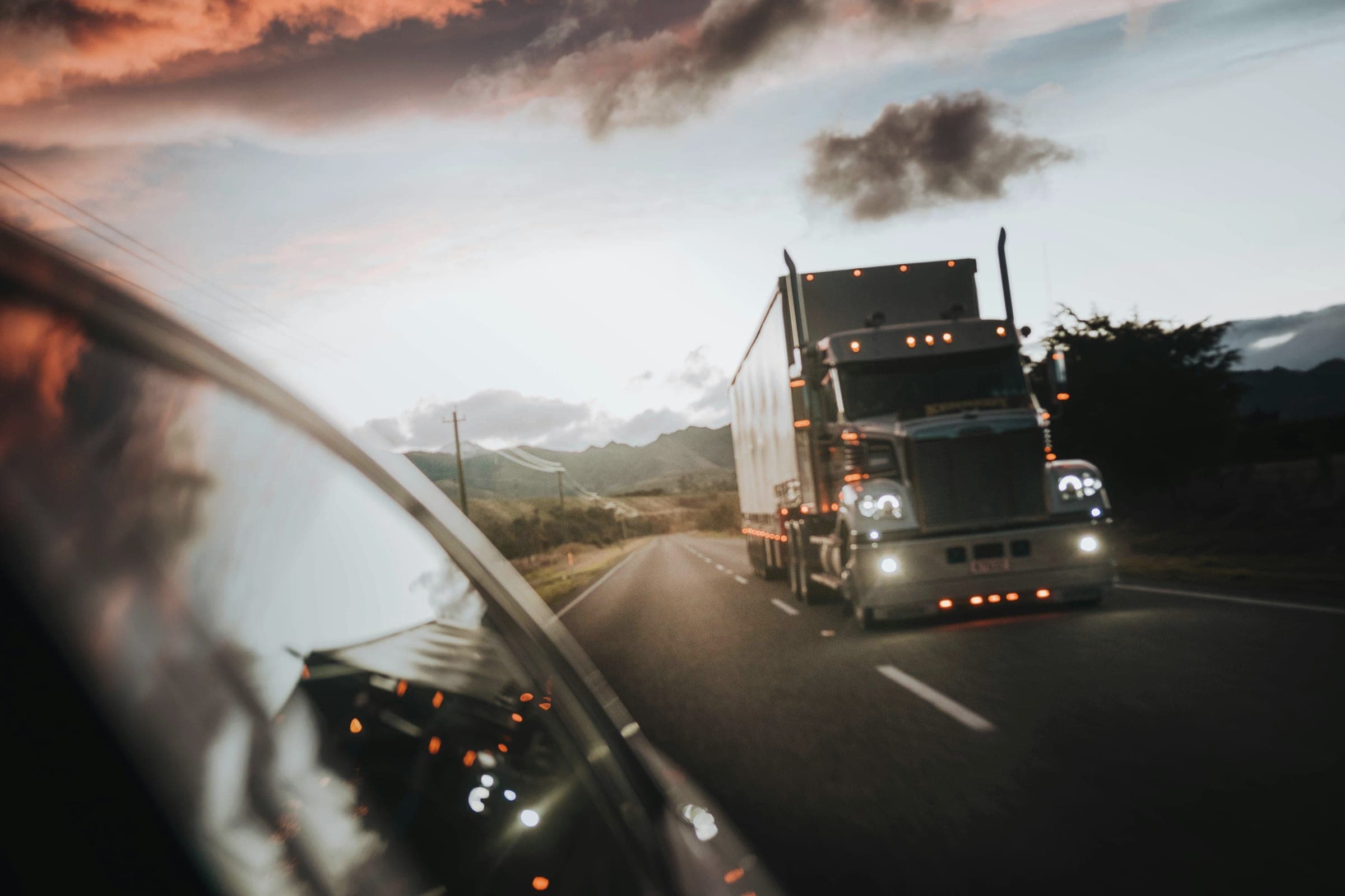 A tractor-trailer truck with its lights on approaching on a mountain road at dusk, seen from a car window.